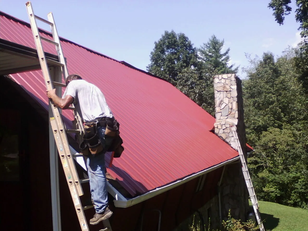 Roofer on a ladder beside a red metal roof and stone chimney — Soffit & Fascia Repair by Roofing Brothers in Stennis Space Center
